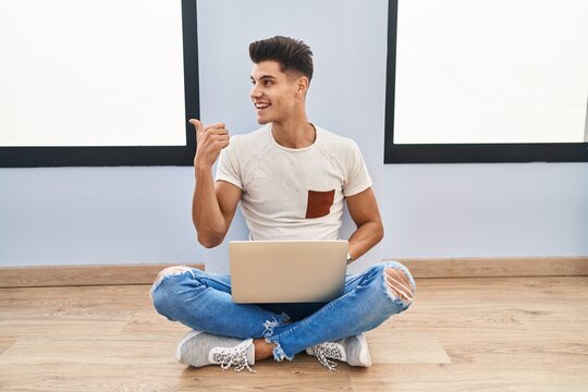 Young hispanic man using laptop at home smiling with happy face looking and pointing to the side with thumb up.