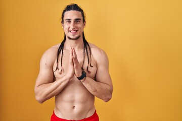 Fototapeta premium Hispanic man with long hair standing shirtless over yellow background praying with hands together asking for forgiveness smiling confident.