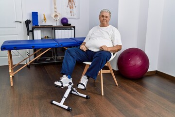 Senior caucasian man at physiotherapy clinic using pedal exerciser winking looking at the camera...