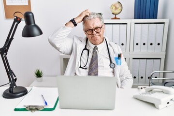 Senior caucasian man wearing doctor uniform and stethoscope at the clinic confuse and wondering about question. uncertain with doubt, thinking with hand on head. pensive concept.