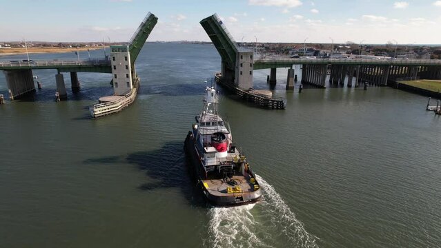 An aerial view behind a tugboat heading inland from the sea. Taken on a sunny day over the East Rockaway Inlet in Queens, NY, the drone camera dolly in following the boat under an opened drawbridge.