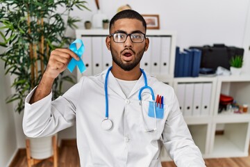 Young indian doctor holding blue ribbon scared and amazed with open mouth for surprise, disbelief...