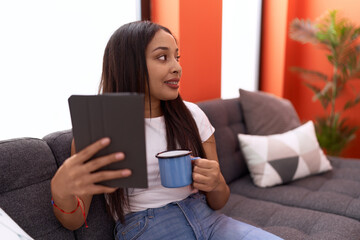 Young arab woman using touchpad drinking coffee sitting on sofa at home