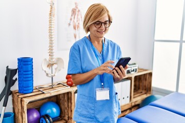 Middle age blonde woman wearing physiotherapist uniform using smartphone at clinic