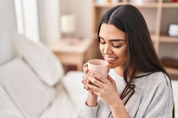 Young hispanic woman drinking cup of coffee sitting on bed at bedroom