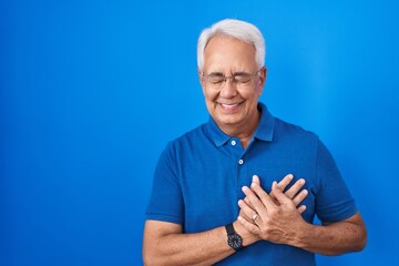Middle age man with grey hair standing over blue background smiling with hands on chest with closed eyes and grateful gesture on face. health concept.