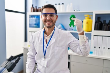 Young hispanic man with beard working at scientist laboratory holding green ribbon looking positive and happy standing and smiling with a confident smile showing teeth