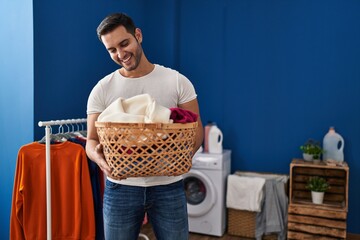 Young hispanic man smiling confident holding basket with clothes at laundry room