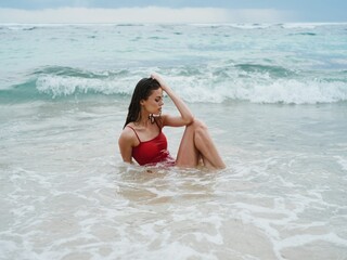 Woman with a beautiful tan tourist in a red swimsuit sitting on the sand on the beach in the ocean in the waves, cloudy weather