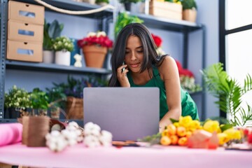 Young hispanic woman florist using laptop talking on smartphone at florist shop