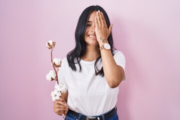 Brunette woman standing over pink background covering one eye with hand, confident smile on face and surprise emotion.