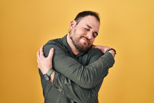Plus size hispanic man with beard standing over yellow background hugging oneself happy and positive, smiling confident. self love and self care
