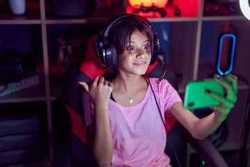 Young girl playing video games with smartphone pointing thumb up to the side smiling happy with open mouth