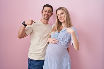 Young couple expecting a baby standing over pink background looking confident with smile on face, pointing oneself with fingers proud and happy.