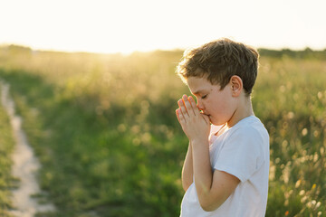 Boy closed her eyes and praying in a field at sunset. Hands folded in prayer concept for faith, spirituality and religion.