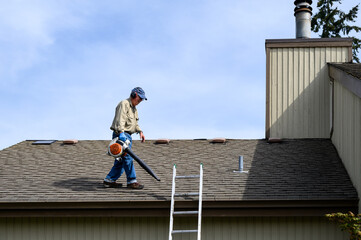 Fall maintenance, senior man on a residential house roof with a gas powered leaf blower cleaning off leaves and pine needles
