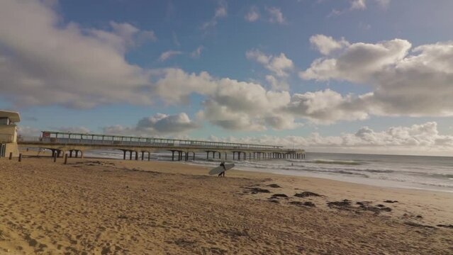 Two surfers with boards, walking on the beach towards group of surfers.Pier in the background, sun creating silhouettes of the group. Slow pan, L-R, wide angle. South Coast UK.