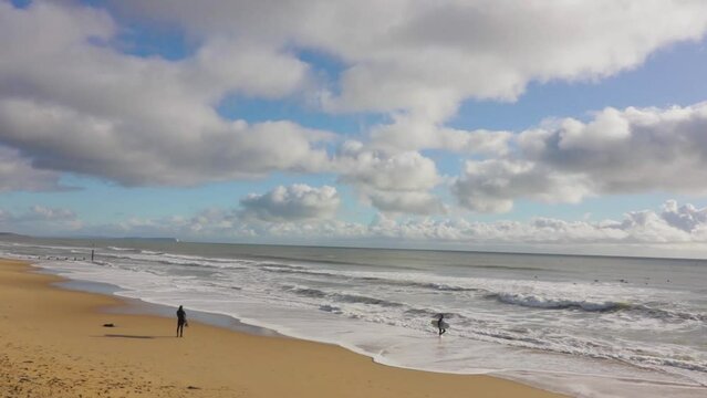 A lone surfer stands watching the waves,  beach and partly cloudy sky in the background.  Stationary wide angle shot. South Coast UK.