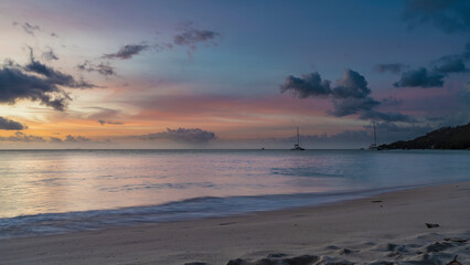 Romantic evening on a tropical beach. Blue clouds in the sky, highlighted in pink, scarlet, orange. Yachts and a hill on the horizon. Foam of waves on the sand. Long exposure. Seychelles. Mahe.