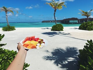 A plate with tropical fruits against the backdrop of a luxurious white beach, turquoise water and palm trees. The concept of holidays in the islands of the Maldives, tourism, travel and vacation.