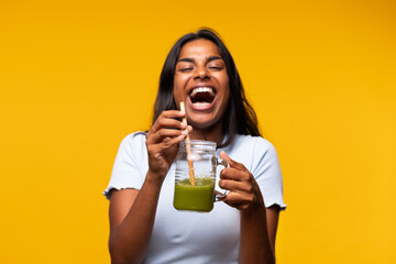 Happy Indian woman with green smoothie on yellow background, laughing. Healthy lifestyle