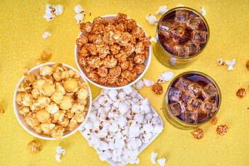 three bowls of spicy, caramelized and salted popcorn accompanied by 2 glasses of soda with ice on a yellow background with copy space for advertising.