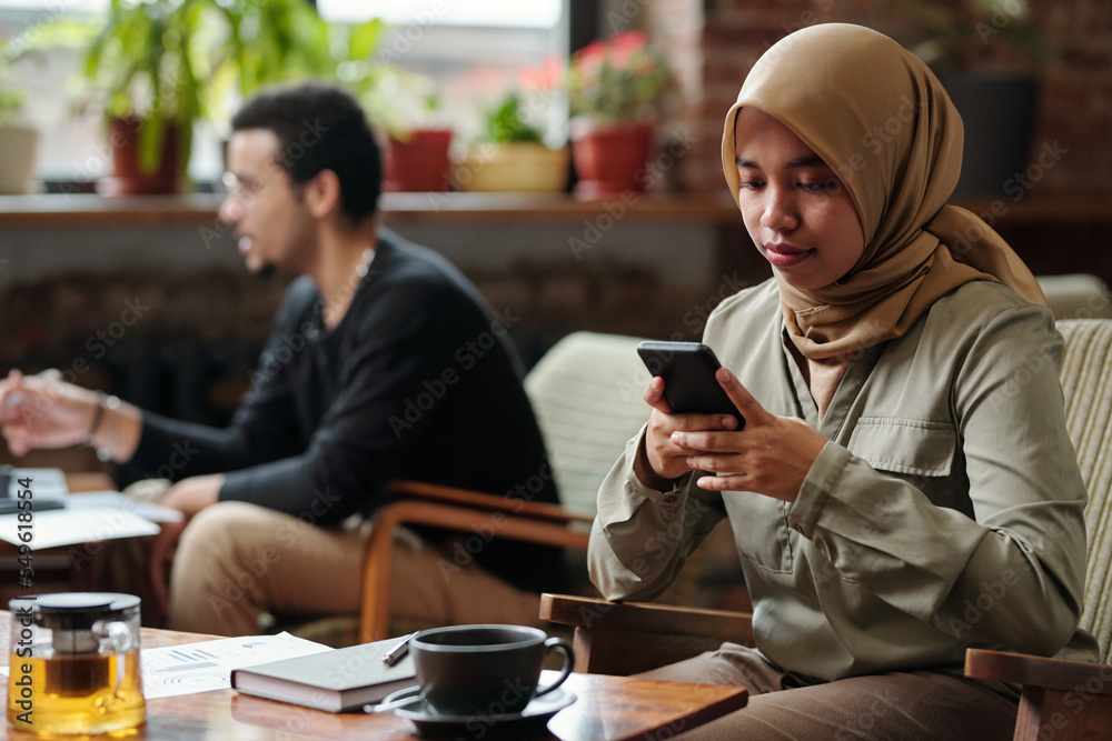 Poster young muslim businesswoman in hijab messaging in smartphone or organizing work while sitting by tabl