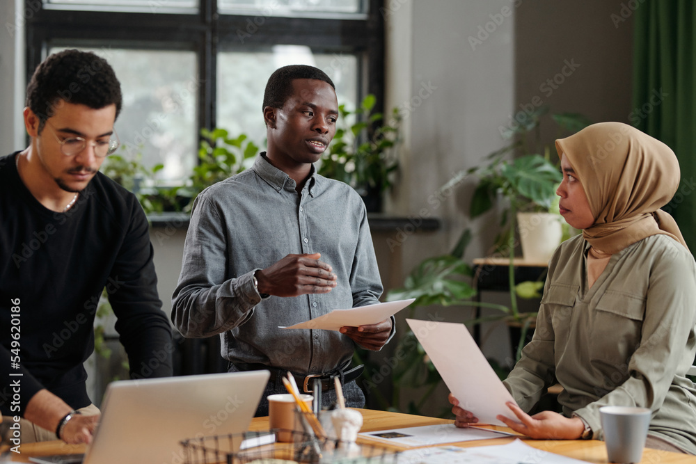 Poster two young intercultural colleagues discussing financial documents by workplace while multi-ethnic ma