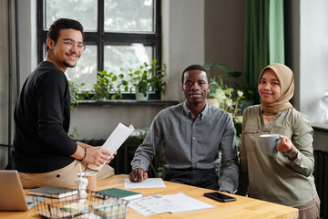 Successful team of young intercultural economists in casualwear looking at camera while sitting by workplace and organizing work at meeting