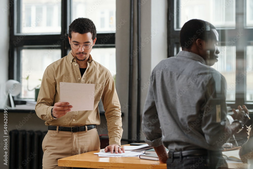 Poster young serious businessman looking through financial document while standing by table in front of afr