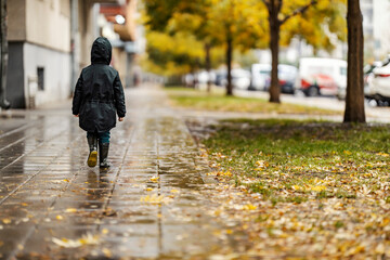 A little boy in raincoat and boots is walking on the city street.