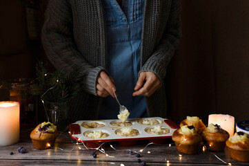 A woman puts muffin dough into a mold in the kitchen. A female chef cooks muffins on a wooden table. Muffin dough with custard and berries. Christmas style.