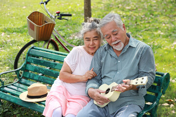 Happy smiling asian senior man and woman sitting on bench playing ukulele and singing a song in...