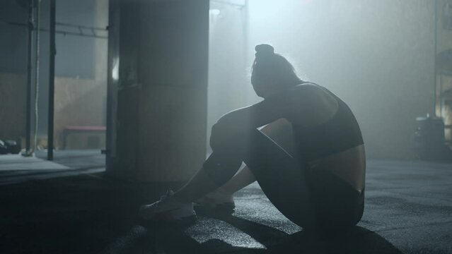 athletic woman is crying after training in gym from fail and pain, sitting on floor alone, rear view