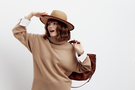 a sweet, happy woman stands on a white background in a beige autumn sweater and holding a bag on her shoulder straightens her hat while looking at toron with a pleasant smile