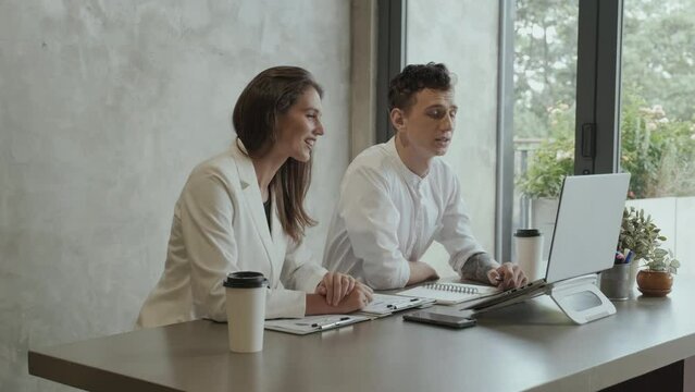 Two young Caucasian coworkers in white formalwear greeting online colleague on laptop screen by waving hand and going on discussion of working points together