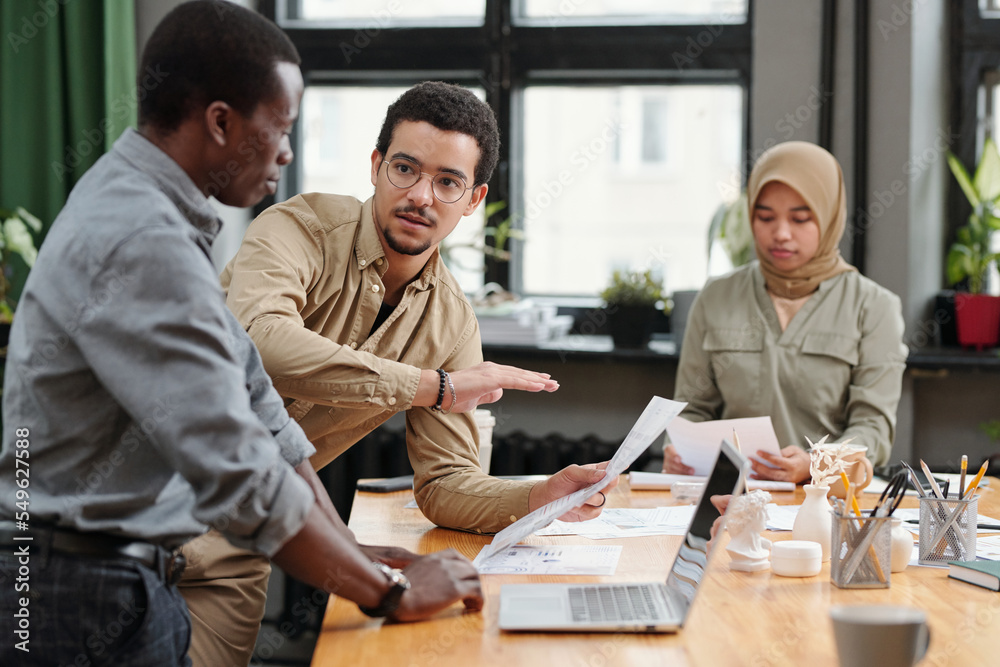 Aufkleber Young confident businessman with document explaining financial rate and results of data analysis while making presentation to colleagues