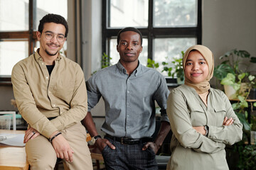 Group of three young multicultural employees in casual attire standing in row in front of camera in openspace office and looking at you
