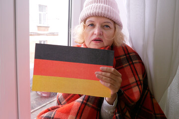 Woman sits in an apartment near window in winter hat and wrapped in a blanket with  cup of hot tea in her hand. Increase price of natural gas for heating in Europe countries. Energy crisis in Europe. 