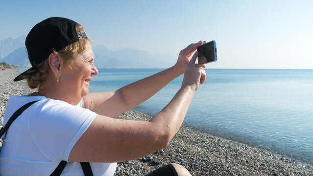 Senior adventureisageless woman with backpack taking a selfie on a mobile phone on the sea beach. Summer active tourism for pensioner. Internet for holiday