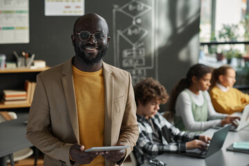 Portrait of African American IT teacher smiling at camera standing in classroom with students using computers in background