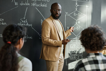 African American teacher explaining formula on blackboard to children during IT technology lesson