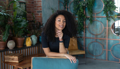 Middle-aged curly black woman in posing over green house plant loft blue background. Looking at camera. Copy space, mock-up. Eco natural, environment conservation positive winter concept.