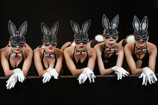 Five young girls of European appearance with brightly painted lips in rabbit masks, bow ties and white gloves stand on all fours leaning on their elbows on stage on a black background.