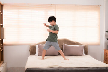 Happy asian elementary school boy jumping and dancing on bed, kid jumped and stomped around joyfully alone in bedroom without feeling tired. little boy have fun on the weekend. healthy child concept.