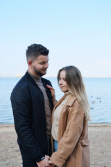 A bearded young man in a black jacket looks at a girl in a coat and holds her hand