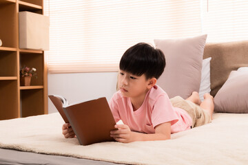 Asian handsome boy reading favorite book while lying down on bed at home, happy little kid spend...