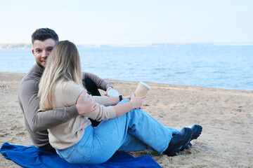 Date on the beach. A young couple in sweaters sits on a plaid and holds stacks of coffee in their hands. The guy hugs the girl and looks at the camera