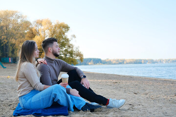 A beautiful young couple, a guy and a girl, are sitting on the beach and drinking coffee. Man and woman enjoy lake view