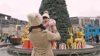 smiling asian Japanese mother raising and playing with her innocent baby girl on a city square with Christmas tree decoration during the winter holiday.